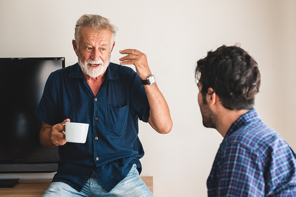 An older man holding a mug, talking to a younger man facing him.